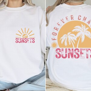 May include: White sweatshirt with a pink and yellow graphic design on the front that says "SUNSETS" and a pink and yellow graphic design on the back that says "FOREVER CHASING SUNSETS" with palm trees and a sun.