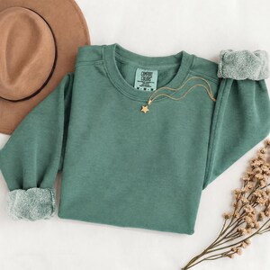 K&ouml;nnte beinhalten: Ein salbeigr&uuml;nes Sweatshirt mit Rundhalsausschnitt, langen &Auml;rmeln und gerippten B&uuml;ndchen, das am inneren Halsausschnitt ein Comfort Colors Markenetikett aufweist. Eine zarte goldfarbene Kette mit einem kleinen sternf&ouml;rmigen Anh&auml;nger liegt auf dem Brustbereich.