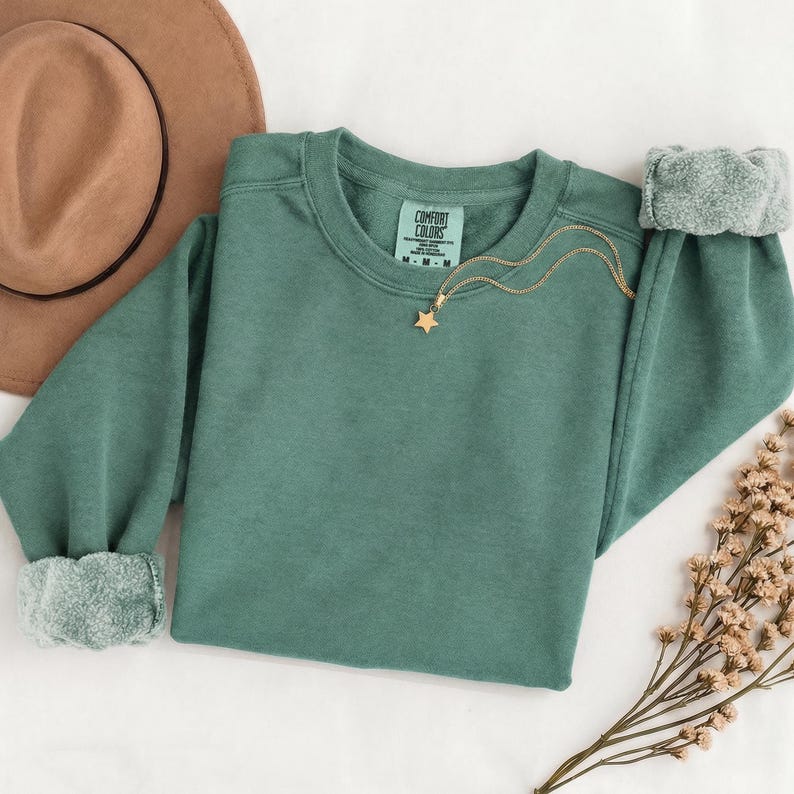 K&ouml;nnte beinhalten: Ein salbeigr&uuml;nes Sweatshirt mit Rundhalsausschnitt, langen &Auml;rmeln und gerippten B&uuml;ndchen, das am inneren Halsausschnitt ein Comfort Colors Markenetikett aufweist. Eine zarte goldfarbene Kette mit einem kleinen sternf&ouml;rmigen Anh&auml;nger liegt auf dem Brustbereich.