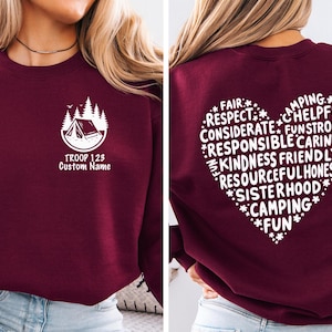 May include: Burgundy sweatshirt with a white camping-themed graphic on the front and a heart-shaped design on the back. The front graphic includes a tent, trees, and the text "TROOP 123 Custom Name." The back features words like "Respect," "Kindness," and "Camping."