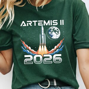 May include: Dark green t-shirt featuring the text "ARTEMIS II 2026" in white. The design includes a rocket launching with flames, a moon, and a starry background. The shirt is a crew neck.