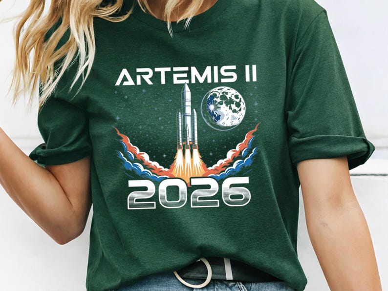 May include: Dark green t-shirt featuring the text "ARTEMIS II 2026" in white. The design includes a rocket launching with flames, a moon, and a starry background. The shirt is a crew neck.
