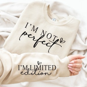 Op de afbeelding: Een beige sweatshirt met de tekst "I'M NOT perfect" in zwart script. De mouw toont "I'M LIMITED edition" in zwart script. De sweatshirt is op een witte ondergrond gevouwen.
