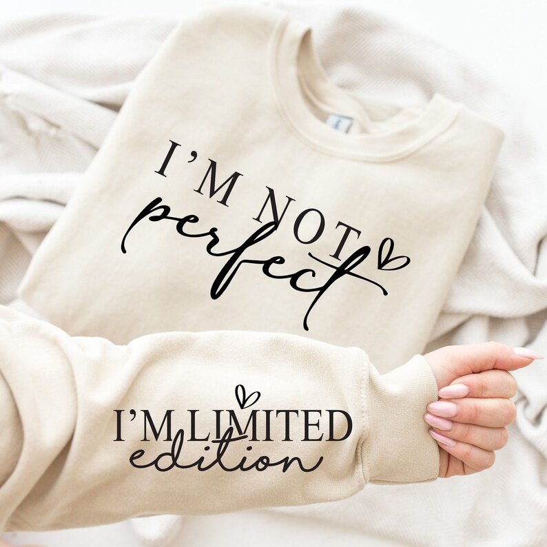 Op de afbeelding: Een beige sweatshirt met de tekst "I'M NOT perfect" in zwart script. De mouw toont "I'M LIMITED edition" in zwart script. De sweatshirt is op een witte ondergrond gevouwen.