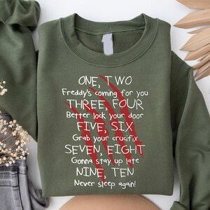 May include: An olive green sweatshirt with the text "ONE, TWO Freddy's coming for you" and other lines from a horror movie. The text is in white, with red claw marks. The sweatshirt is folded, with a sleeve visible.