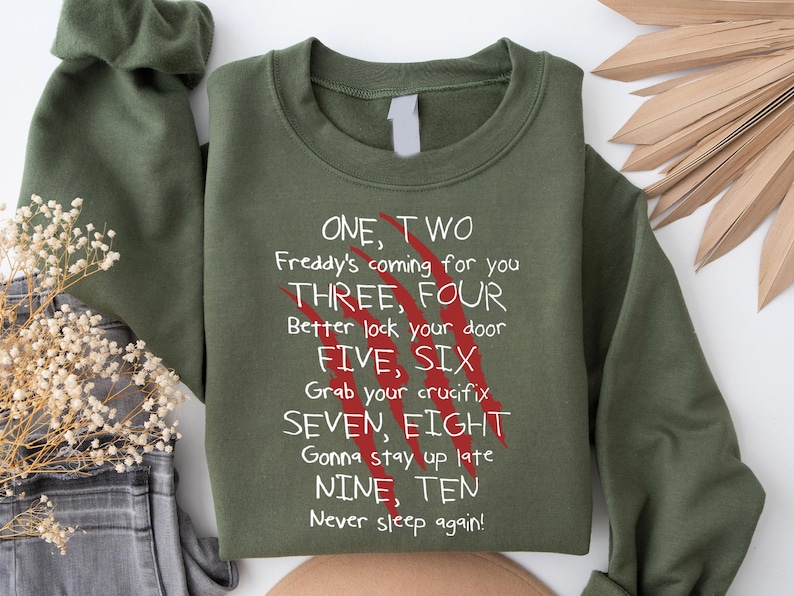 May include: An olive green sweatshirt with the text "ONE, TWO Freddy's coming for you" and other lines from a horror movie. The text is in white, with red claw marks. The sweatshirt is folded, with a sleeve visible.