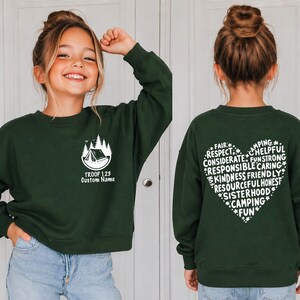May include: A dark green sweatshirt with a white camping graphic and text. The front features a tent and trees with "TROOP 123 Custom Name". The back displays a heart-shaped design with words like "Fair", "Respect", and "Camping".