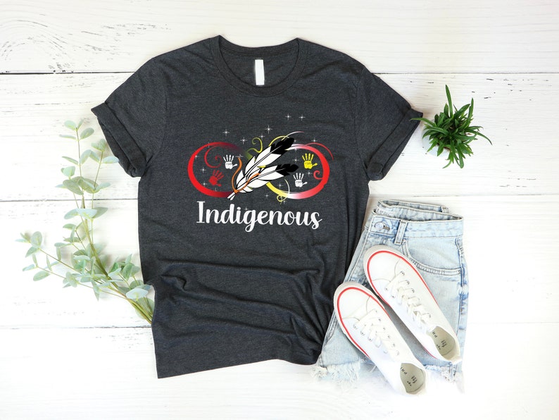May include: A dark grey t-shirt with an infinity symbol design featuring a feather and handprints in red, yellow, orange, and black. The text "Indigenous" is printed below the symbol.