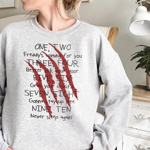 May include: A light gray sweatshirt with the text "ONE, TWO Freddy's coming for you THREE, FOUR Better lock your door FIVE SIX Grab your crucifix SEVEN, EIGHT Gonna stay up late NINE TEN Never sleep again!" and red claw marks.