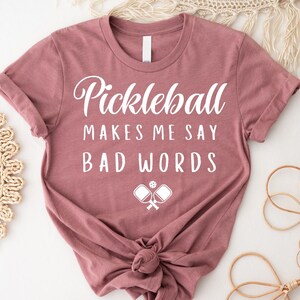 May include: A mauve t-shirt with the text "Pickleball Makes Me Say Bad Words" in white lettering. A white silhouette of two pickleball paddles and a ball is below the text.