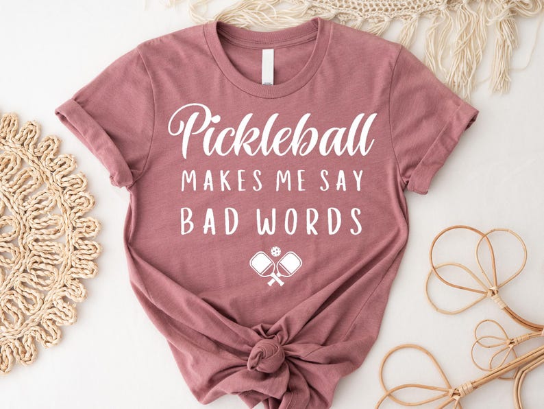 May include: A mauve t-shirt with the text "Pickleball Makes Me Say Bad Words" in white lettering. A white silhouette of two pickleball paddles and a ball is below the text.