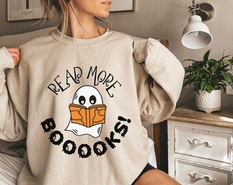 May include: A beige crewneck sweatshirt featuring a graphic of a white ghost reading an orange book with black stitching details. The text above the ghost reads READ MORE and the text below the ghost reads BOOOOKS! in a black, dripping font.