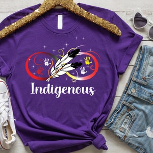 May include: Purple t-shirt with an infinity symbol design featuring a feather and handprints in red, yellow, and blue. The word "Indigenous" is printed below the symbol.