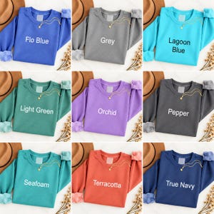 K&ouml;nnte beinhalten: Ein Raster aus neun lang&auml;rmeligen Sweatshirts mit Rundhalsausschnitt in verschiedenen Farben wie Flo Blue, Grey, Lagoon Blue, Light Green, Orchid, Pepper, Seafoam, Terracotta und True Navy. Jedes Sweatshirt hat eine goldene Sternkette.