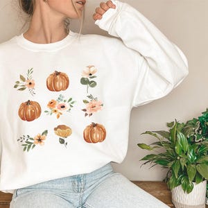 Puede incluir: Sudadera blanca con un dise&ntilde;o de acuarela de calabazas y detalles florales en tonos naranja, melocot&oacute;n y verde. El dise&ntilde;o se extiende por la parte delantera de la parte superior de manga larga, creando una est&eacute;tica oto&ntilde;al.