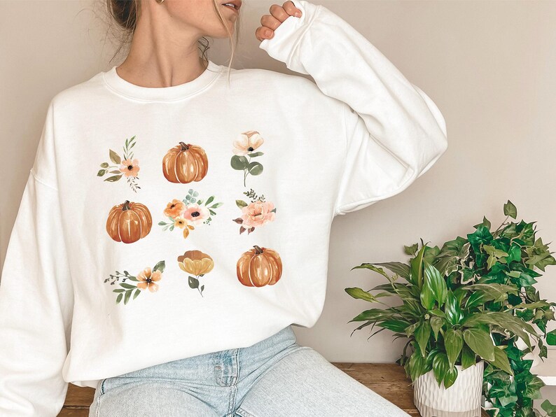 Puede incluir: Sudadera blanca con un dise&ntilde;o de acuarela de calabazas y detalles florales en tonos naranja, melocot&oacute;n y verde. El dise&ntilde;o se extiende por la parte delantera de la parte superior de manga larga, creando una est&eacute;tica oto&ntilde;al.