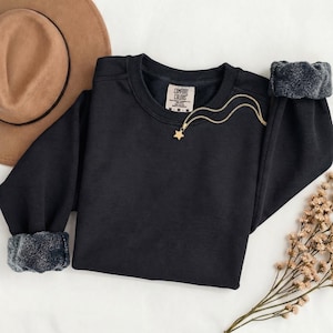 K&ouml;nnte beinhalten: Ein schwarzes Langarm-Sweatshirt mit Rundhalsausschnitt und einem Comfort Colors Markenetikett am Innenkragen. Das Sweatshirt verf&uuml;gt &uuml;ber gerippte B&uuml;ndchen und eine goldfarbene Halskette mit einem kleinen Sternanh&auml;nger auf dem Stoff.