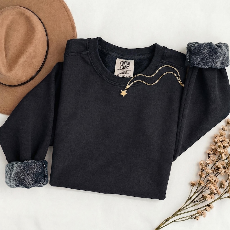 K&ouml;nnte beinhalten: Ein schwarzes Langarm-Sweatshirt mit Rundhalsausschnitt und einem Comfort Colors Markenetikett am Innenkragen. Das Sweatshirt verf&uuml;gt &uuml;ber gerippte B&uuml;ndchen und eine goldfarbene Halskette mit einem kleinen Sternanh&auml;nger auf dem Stoff.