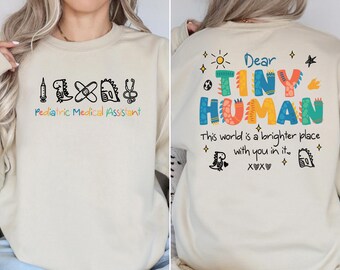 May include: A cream-colored sweatshirt with a graphic design on the front that says "Pediatric Medical Assistant" with a syringe, dinosaur, bandage, and stethoscope. The back of the sweatshirt has a graphic design that says "Dear Tiny Human. This world is a brighter place with you in it. XXX" with a dinosaur, heart, and stars.