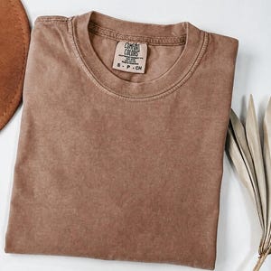 May include: A solid brown short-sleeve t-shirt with a crew neckline. The interior neck label features the text Comfort Colors, 100% Cotton, Made in Honduras, and size S-P-CH. The fabric has a slightly faded, washed-out appearance.