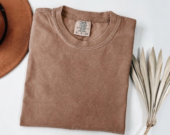 Könnte beinhalten: Ein einfarbig braunes T-Shirt mit kurzen Ärmeln und Rundhalsausschnitt. Das Etikett im Nackenbereich zeigt den Text Comfort Colors, 100% Cotton, Made in Honduras sowie die Größe S-P-CH. Der Stoff hat eine leicht verwaschene Optik.