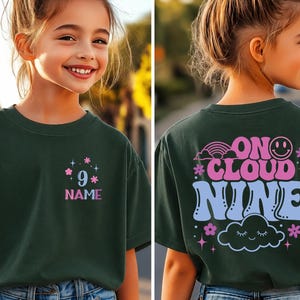 May include: Dark green t-shirt with the text "9" and "NAME" in pink and blue, with floral accents. The back of the shirt features the phrase "ON CLOUD NINE" with a rainbow, smiley face, cloud, and floral designs.