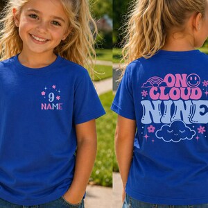 May include: Royal blue t-shirt with a front graphic featuring the number 9, stars, and the word "NAME" in pink and blue. The back of the shirt displays the phrase "ON CLOUD NINE" with a rainbow, smiley face, cloud, and stars.