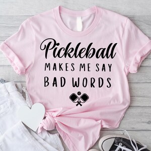 May include: Pink t-shirt with the text "Pickleball makes me say bad words" and a black and white illustration of two pickleball paddles.