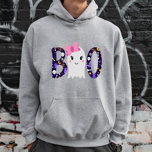 May include: A light gray hooded sweatshirt featuring a graphic on the chest with the text BOO. The letters B and O are filled with a dark purple pattern of skulls, stars, and moons. A white ghost with a pink bow replaces the middle letter O.