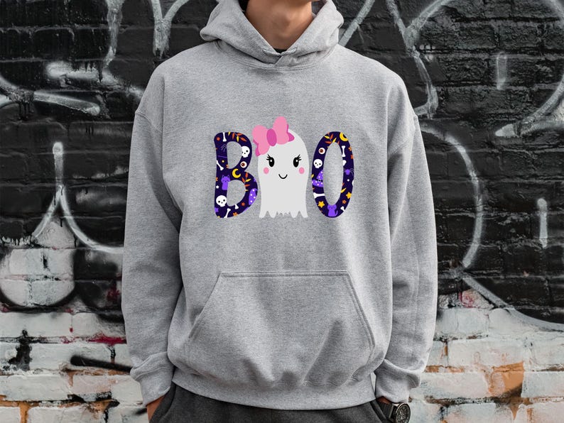 May include: A light gray hooded sweatshirt featuring a graphic on the chest with the text BOO. The letters B and O are filled with a dark purple pattern of skulls, stars, and moons. A white ghost with a pink bow replaces the middle letter O.
