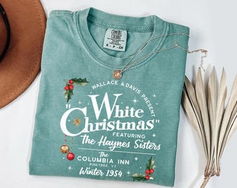 May include: A teal-colored t-shirt with the text "White Christmas" in a vintage-style design. The shirt features holly and ornaments, with the text "The Haynes Sisters" and "Winter 1954". A gold necklace and a brown hat are also visible.