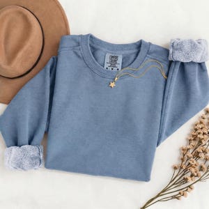 K&ouml;nnte beinhalten: Ein blaues Sweatshirt mit Rundhalsausschnitt, langen &Auml;rmeln und gerippten B&uuml;ndchen, das am inneren Halsausschnitt ein Comfort Colors Markenetikett aufweist. Eine goldfarbene Kette mit einem kleinen sternf&ouml;rmigen Anh&auml;nger liegt auf dem Kleidungsst&uuml;ck.