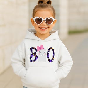 May include: A white hooded sweatshirt featuring a graphic print on the front. The design displays the word BOO in dark purple with a pattern of skulls, stars, and moons. The letter O is replaced by a white ghost wearing a pink bow.