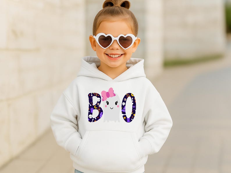 May include: A white hooded sweatshirt featuring a graphic print on the front. The design displays the word BOO in dark purple with a pattern of skulls, stars, and moons. The letter O is replaced by a white ghost wearing a pink bow.