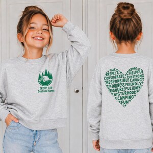 May include: A light gray sweatshirt with a green camping-themed graphic on the front and a heart-shaped design on the back. The front graphic includes a tent, trees, and the text "TROOP 128 Custom Name." The back features words like "Fair," "Respect," and "Camping."