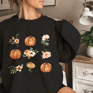 Puede incluir: Sudadera negra con ilustraciones en acuarela de calabazas y arreglos florales en tonos naranja, melocot&oacute;n y crema. El dise&ntilde;o es un patr&oacute;n con tem&aacute;tica oto&ntilde;al, perfecto para el oto&ntilde;o.