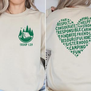 May include: A beige sweatshirt with a green camping graphic and the text "TROOP 123" on the front. The back features a green heart-shaped design with words like "FAIR", "RESPECT", and "CAMPING".
