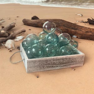 May include: A collection of translucent, spherical, sea-green glass floats piled inside a square, weathered white wooden crate with a rope handle. The glass spheres vary slightly in size and clarity, reflecting light across their smooth surfaces.