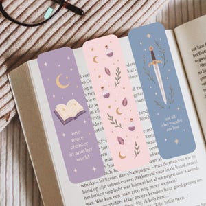 May include: Three decorative bookmarks with celestial and fantasy themes. One is lavender with a crescent moon and open book, another is pink with potion bottles and crystals, and the last is blue with a sword and text.