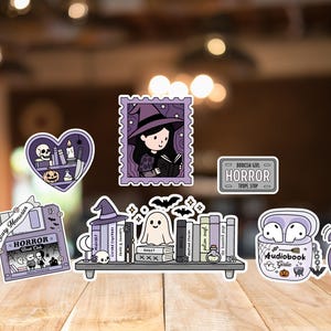 May include: A selection of Halloween-themed stickers. Designs include a witch reading, a ghost, and the word "HORROR". The stickers are in purple, black, and white, with a cartoon style. Measurements are in cm.