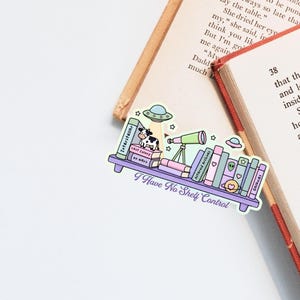 Op de afbeelding: Een kleurrijke sticker met een boekenplankontwerp met boeken, een koe, een telescoop en UFO's. De tekst "I Have No Shelf Control" staat op de sticker, die op een open boek ligt.