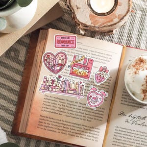 May include: An open book with several pink stickers featuring romantic themes. Stickers include a heart with candles, a shelf of books, and a pink floppy disk with the words "Slow Burn". A cup of coffee sits nearby.