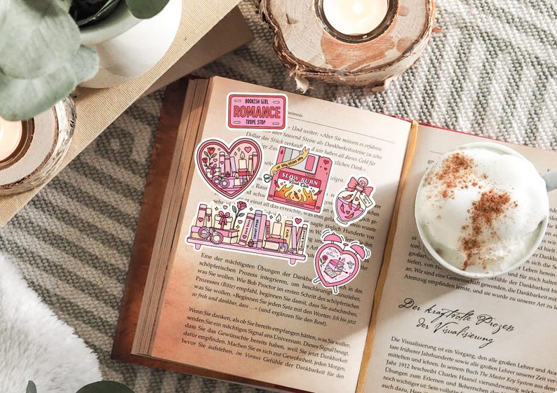 May include: An open book with several pink stickers featuring romantic themes. Stickers include a heart with candles, a shelf of books, and a pink floppy disk with the words "Slow Burn". A cup of coffee sits nearby.