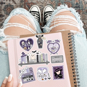 May include: A pink notebook with spiral binding and Halloween stickers. The stickers depict a coffin, a witch, a ghost, and the word "HORROR". The person is wearing ripped blue jeans and black and white trainers.