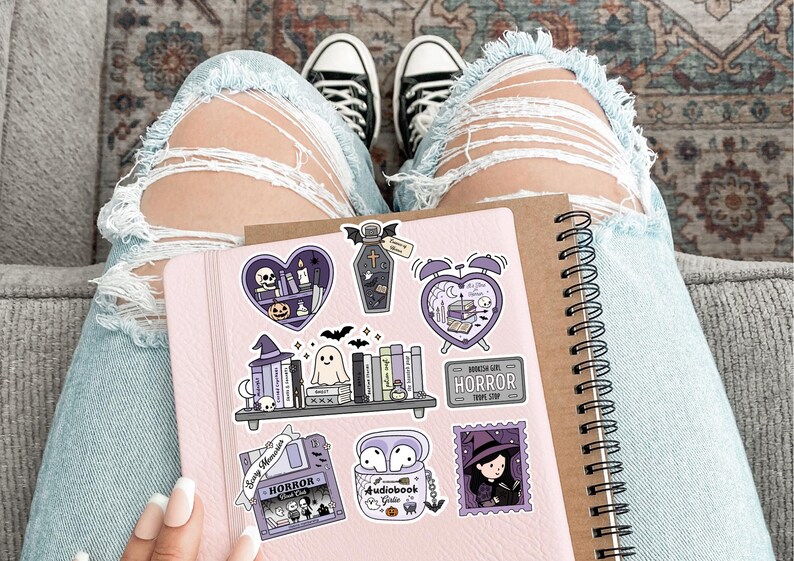May include: A pink notebook with spiral binding and Halloween stickers. The stickers depict a coffin, a witch, a ghost, and the word "HORROR". The person is wearing ripped blue jeans and black and white trainers.