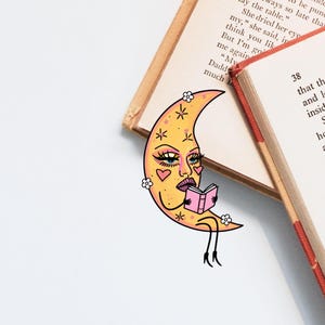 May include: A whimsical bookmark featuring a yellow crescent moon with a face, reading a pink book. The bookmark is decorated with hearts and flowers. Two open books are in the background.