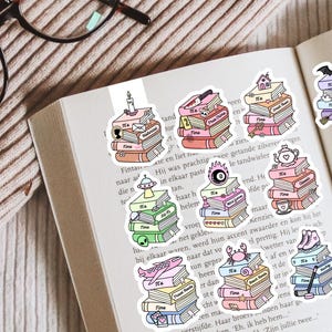 May include: A collection of ten die-cut stickers featuring stacks of books with various thematic icons on top, including a candle, knife, house, bat, UFO, magic eight ball, teapot, dragon, aeroplane, crab, ice skates, and cowboy boots.