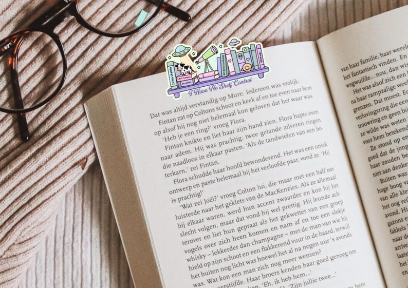 Op de afbeelding: Een kleurrijke boekenlegger met een plankontwerp, boeken, een telescoop en een koe in een ruimteschip. De tekst "I Have No Shelf Control" is zichtbaar. De boekenlegger ligt in een open boek.