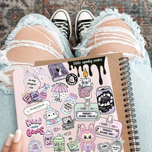 May include: A spiral-bound notebook and a pink sticker sheet with various spooky-themed illustrations and text, including "little spooky reader" and "Read More Books." The person is wearing light blue ripped jeans and black and white sneakers.