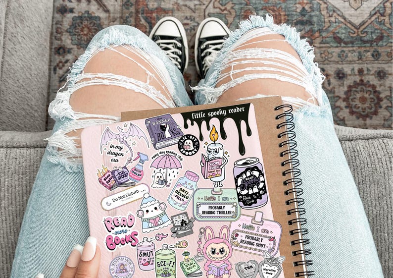 May include: A spiral-bound notebook and a pink sticker sheet with various spooky-themed illustrations and text, including "little spooky reader" and "Read More Books." The person is wearing light blue ripped jeans and black and white sneakers.